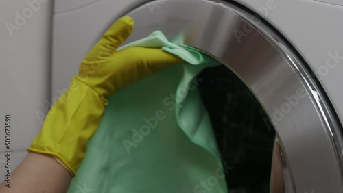 Housewife is engaged in wet cleaning in bathroom wipes washing machine from moisture so that there is no mold. Girl in yellow protective gloves wipes door of washing machine dryly with a green napkin photo