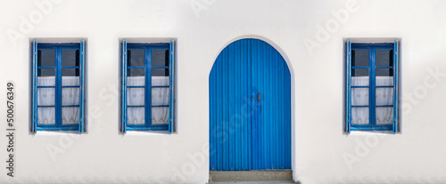 Blue door and three windows with open shutters on white wall. Greek island house front view