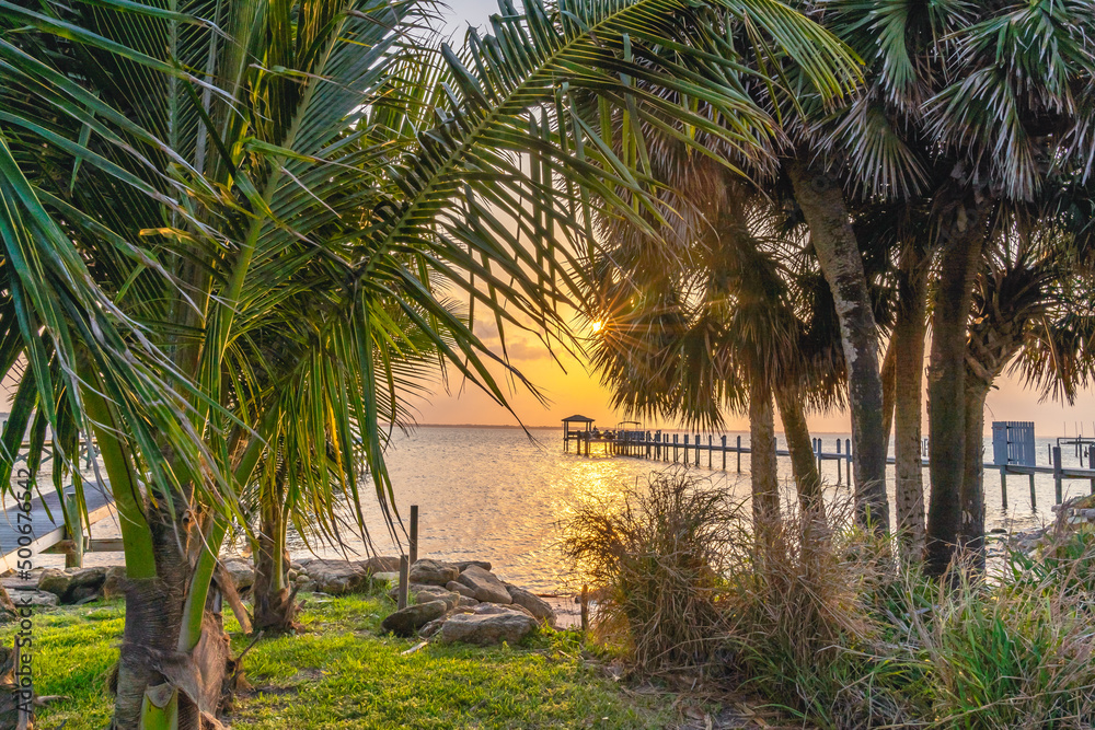 View of a yellow sunset over the Indian River through a gap between palm trees from the A1A in Florida. Typical wooden pier for parking boats in shallow water
