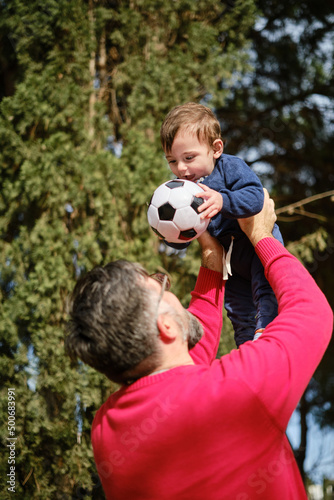 Man having fun with his little son while holding him up in the air with a soccer ball. © alvaro
