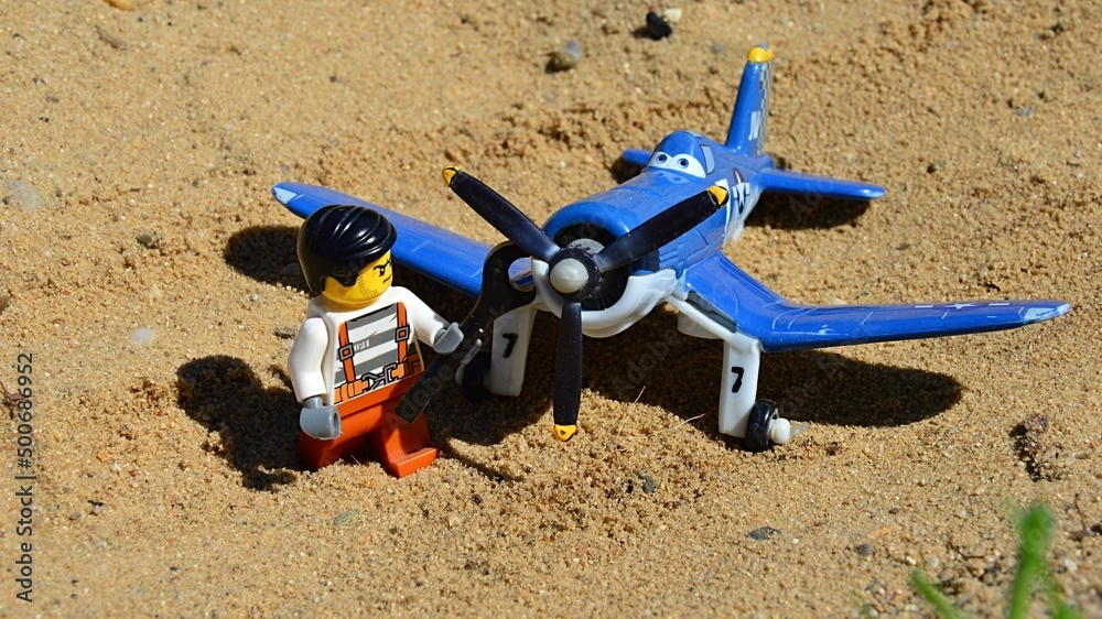 LEGO City figure of gloomy prisoner is tightening propeller of Vought F4U Corsair aircraft model named Skipper from Disney Pixar animated movie Planes on sandy sunlit ground. Photo | Adobe Stock