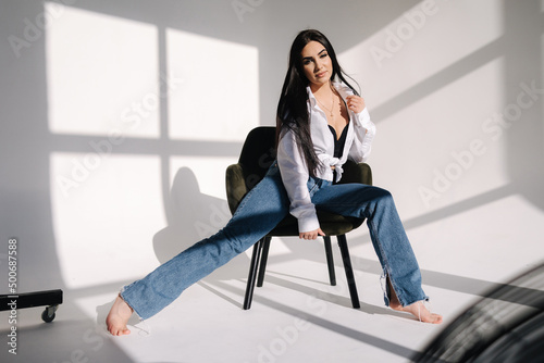 Pretty woman has photosession in studio. Sexy girl in denim and white shirt. Barefoot woman. Light from the sun makes a shadow on a white background. Stylish green chair