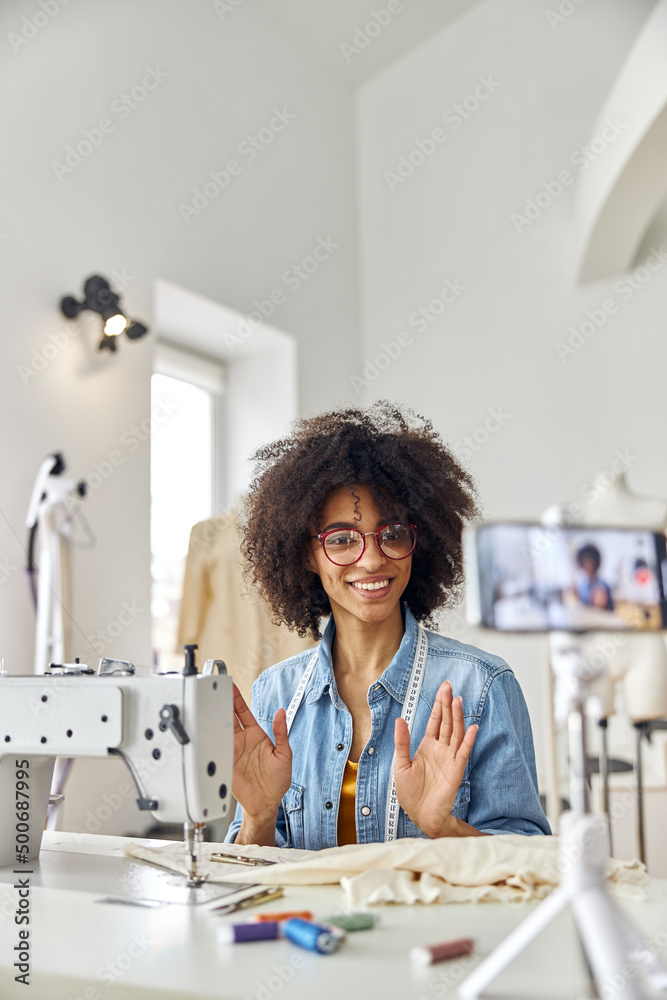 African-American woman dressmaker looks at phone camera at workplace with sewing machine in studio African-American woman dressmaker looks at phone camera at workplace with sewing machine in studio