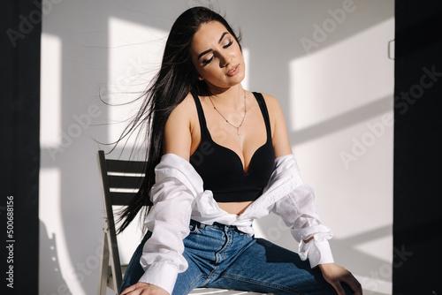 Sexy woman in black topic and blue jeans sits on chair during studio photoshoots. Brunette girl posing to photographer. Shadow on white background