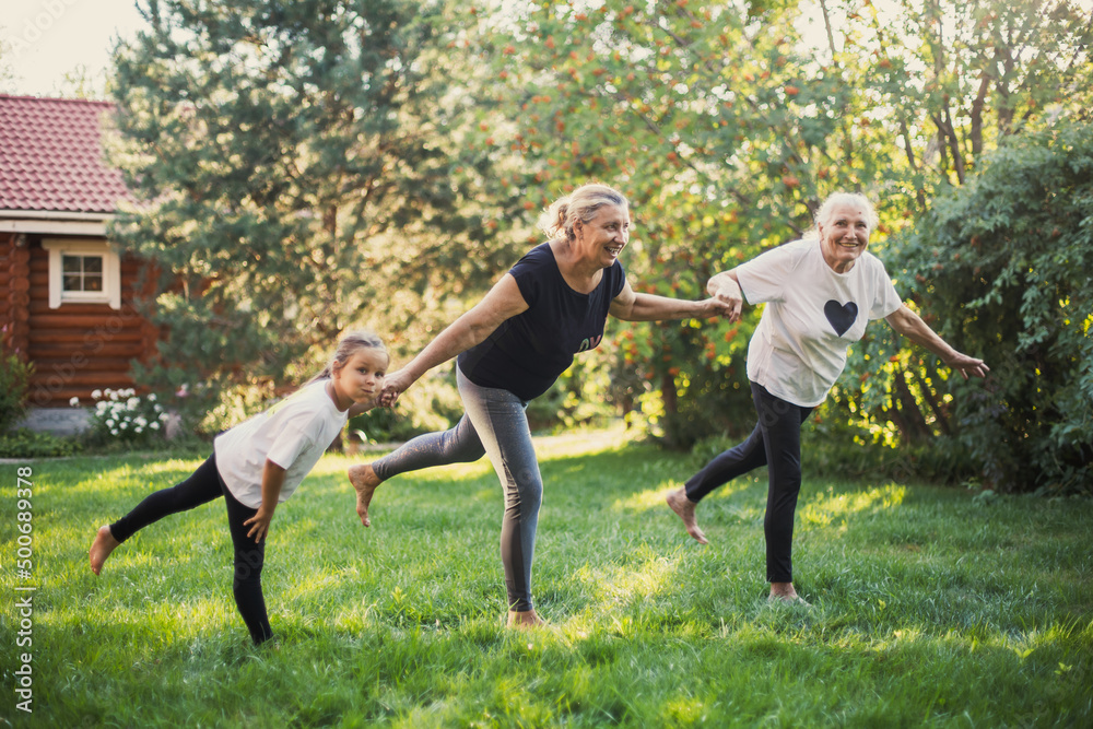 Three generations of family females smiling balancing on one leg, holding hands doing physical exercises together on backyard on meadow full of green grass and trees. Spending time together. Three generations of family females smiling balancing on one leg, holding hands doing physical exercises together on backyard on meadow full of green grass and trees. Spending time together.