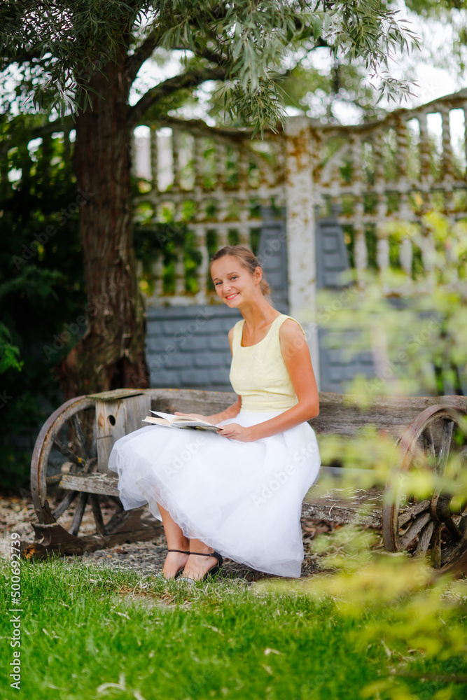 Cute european girl woman with a book on a garden bench under a willow tree, a beautiful garden and a wooden bench made of wagon wheels, landscape art and a beautiful well-groomed garden Cute european girl woman with a book on a garden bench under a willow tree, a beautiful garden and a wooden bench made of wagon wheels, landscape art and a beautiful well-groomed garden