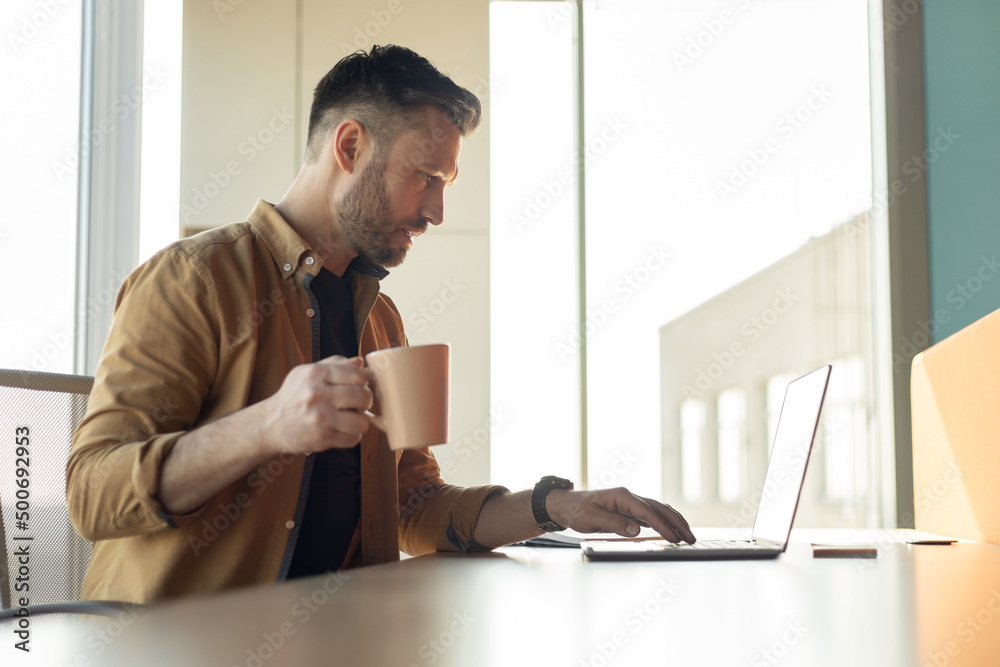 Man Working On Laptop Drinking Coffee In Office, Side View