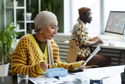 Side view portrait of Muslim young woman wearing headscarf and eating healthy lunch in office photo