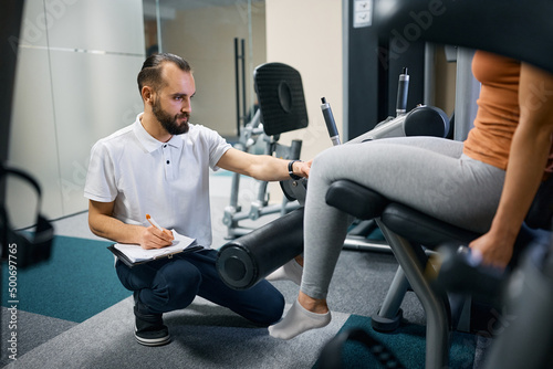 Physiotherapist taking notes while assisting sportswoman in exercising on leg extension machine at health club.