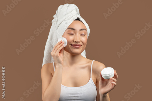 Happy pretty asian woman cleansing her skin with cotton pad, holding jar with cream and smiling over brown background