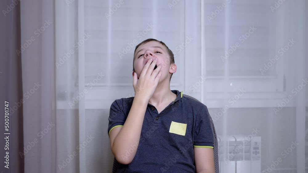 Sleepy Child Yawns, Covers his Mouth with his Hand, Sitting in a Chair in Room. A smiling, bored, tired boy in a t-shirt opens his mouth wide. Change in facial expression. Emotions. Gesture. Bedtime. Sleepy Child Yawns, Covers his Mouth with his Hand, Sitting in a Chair in Room. A smiling, bored, tired boy in a t-shirt opens his mouth wide. Change in facial expression. Emotions. Gesture. Bedtime.