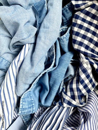 Background photo of dozen of variety of blue colored shirts. Blue fabric, shirt mess. Light to dark blue. Stripped, checkered, jean, cotton textiles in laundry basket. Shades of blue