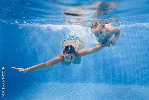 Mom and daughter are immersed in water, swimming under water in a paddling pool. Diving kid. Learning child to swim. Young mother or swimming instructor and happy little girl.