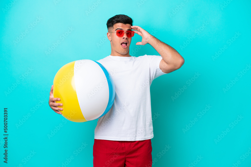 Fototapeta premium Young caucasian man holding a beach ball isolated on blue background doing surprise gesture while looking to the side