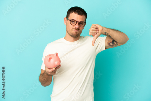 Brazilian man holding a piggybank over isolated blue background showing thumb down with negative expression
