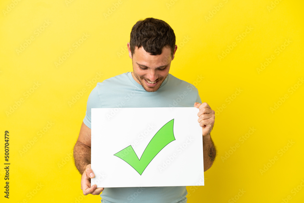 Brazilian man over isolated purple background holding a placard with text Green check mark icon Brazilian man over isolated purple background holding a placard with text Green check mark icon
