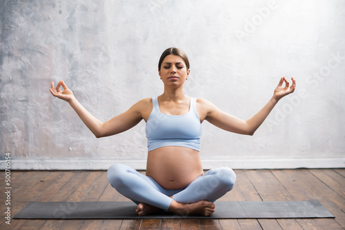 Young pregnant woman doing yoga exercises and meditating at home. Health care, mindfulness, relaxation and wellness concept.