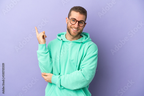 Young handsome caucasian man isolated on purple background happy and pointing up © luismolinero