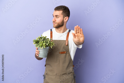 Gardener caucasian man holding a plant isolated on yellow background making stop gesture and disappointed