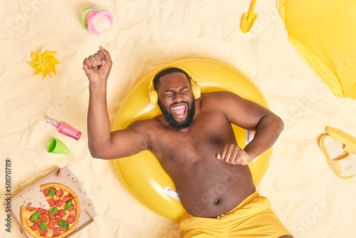 Canvas Print Joyful handsome adult man with dark skin thick beard dances with rhythm of music wears stereo headphones on ears has rest on beach lies in sun has good mood