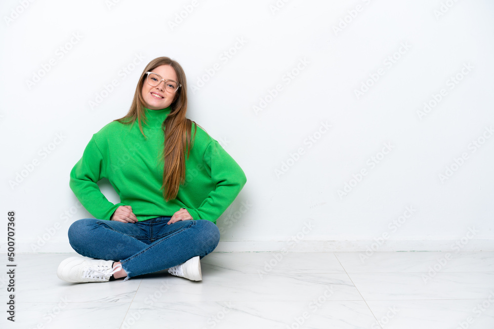 Young caucasian woman sitting on the floor isolated on white background posing with arms at hip and smiling