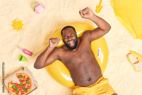 Overhead view of overjoyed dark skinned adult man laughs and shakes arms poses with bare torso has fun on beach lies on inflated yellow swimring eats pizza enjoys lazy day during summer holidays photo