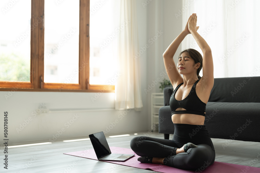 Calm relaxed young asian woman sitting in lotus position with hands up namaste gesture, closed eyes, breathing, resting, deeply meditating, practicing yoga on yoga mat at home. Calm relaxed young asian woman sitting in lotus position with hands up namaste gesture, closed eyes, breathing, resting, deeply meditating, practicing yoga on yoga mat at home.