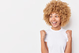 Positive excited curly woman makes winning gesture clenches fists celebrates personal achievements concentrated away wears casual basic t shirt isolated over white background copy space for text