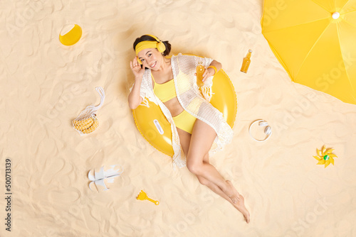 Overhead shot of positive attractive Asian woman dressed in yellow swim suit enjoys listening music poses on inflated swimring drinks energetic drink chills at sandy beach has summer vacation.