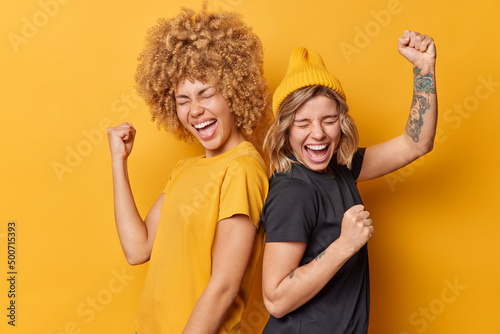 Two positive women friends feel very happy like winners make triumph gesture shake arms celebrate success stand back to each other dressed in casual t shirts isolated over yellow background Fototapet