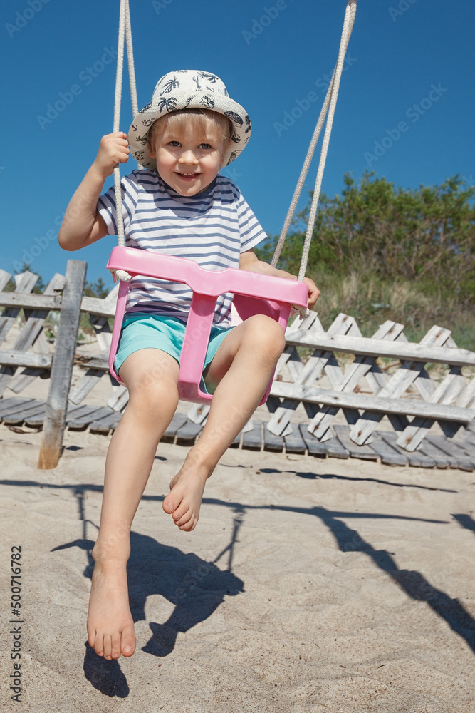 The little hilarious boy swings in a pink swing, he looks straight at the camera and poses. Beach background, sand and blue sky. The little hilarious boy swings in a pink swing, he looks straight at the camera and poses. Beach background, sand and blue sky.