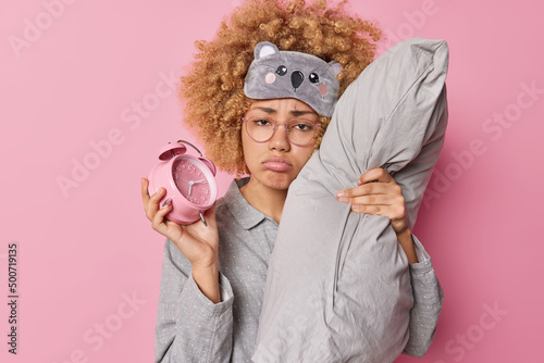 Upset curly haired woman feels sad awakes very early wants to sleep dressed in pajama has bad mood leans on pillow holds alarm clock isolated over pink background. Time to wake up and go for work. photo