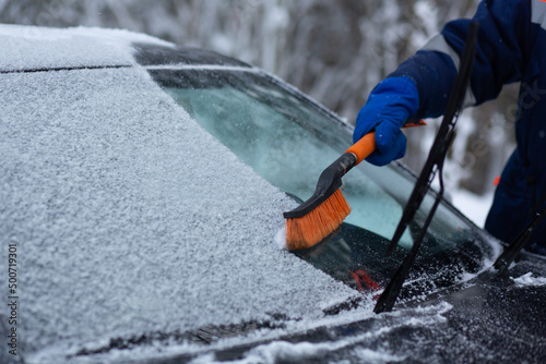 A human hand in a glove cleans a blue car from snow with a special brush. A hand sweeps snow from the hood of the car. Horizontal photo. the consequences of a blizzard