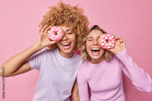 Waist up shot of positive young friendly women cover eye with delicious doughnuts have fun enjoy eating sweet dessert stand closely to each other isolated over pink background. Tasty donuts.