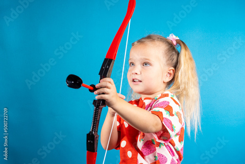 Inspirational girl in a red dress shoots a bow. The concept of children's games, striving for a goal, successfully solving a problem, love and cupid, carefree happy childhood.