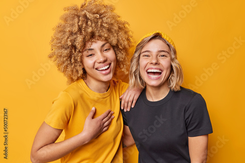 Best female friends laugh happily feel amused smile broadly have fun together dressed in casual t shirts isolated over vivid yellow studio background. Positive human emotions and feelings concept