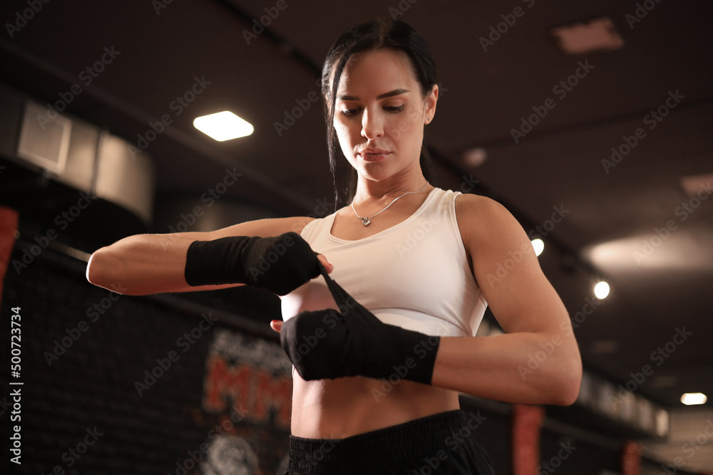 Young woman wraps her fists in black bandages for kickboxing gloves against a black boxing hall