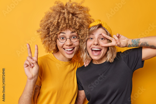 Positive two female friends show peace gesture smile gladfully laugh happily dressed in casual t shirts isolated over yellow background. Playful girls go crazy have good mood make victory sign photo