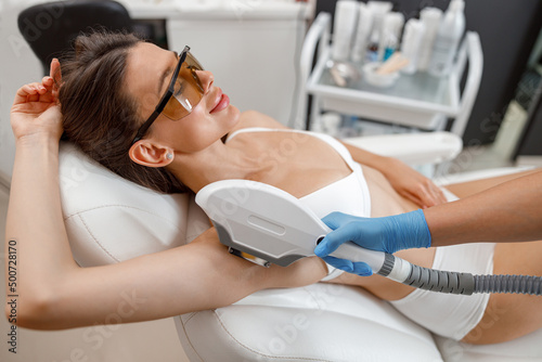 Female client in eyeglasses during armpit photo epilation procedure with ipl machine in beauty salon. photo
