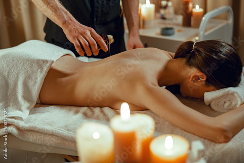 Masseur man therapist pouring warm herb infused oil on female back.