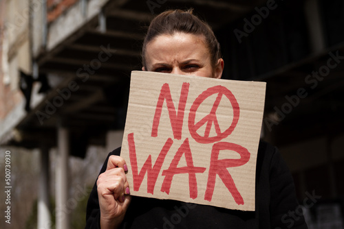 Woman holding "No War" placard with peace sign in protest manifestation against war. Girl in ruined, bombed residential house, abandoned destroyed building. Female with banner, anti-war demonstration.