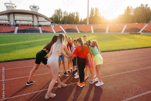 A large group of children, boys and girls, stand together in a circle and fold their hands, tuning up and raising team spirit before the game at the stadium during sunset. A healthy lifestyle. © Andrii