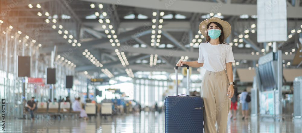 Fototapeta premium Young female wearing face mask with luggage walking in airport, protection Coronavirus disease infection, Asian woman traveler with hat. Time to travel after vaccine booster dose concept