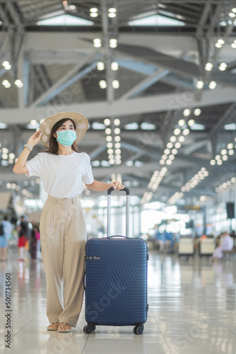 Young female wearing face mask with luggage walking in airport, protection Coronavirus disease infection, Asian woman traveler with hat. Time to travel after vaccine booster dose concept