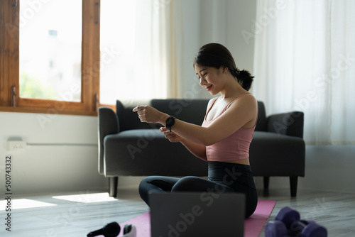 Attractive young asian fitness woman in sportswear resting after workout using smartwatch for online training at home