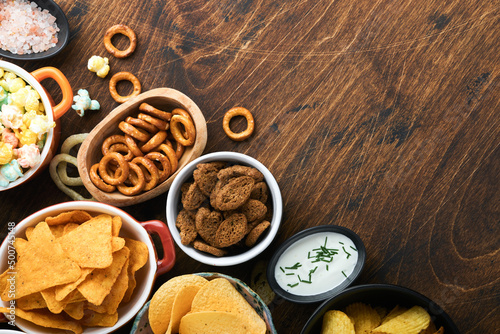 Unhealthy food or snacks. All classic potato snacks with peanuts, popcorn and onion rings and salted pretzels in bowl plates on old wooden background. Unhealthy food for figure, heart, skin, teeth.