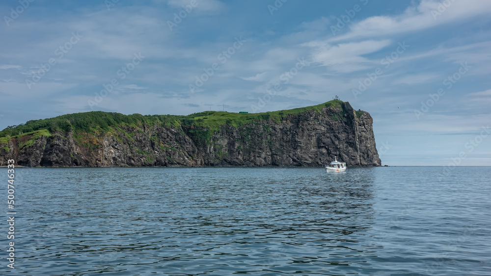 The picturesque coast of Kamchatka against the blue sky. Steep rocky slopes and green vegetation on the hills. A white yacht is sailing on the Pacific Ocean. Avacha Bay