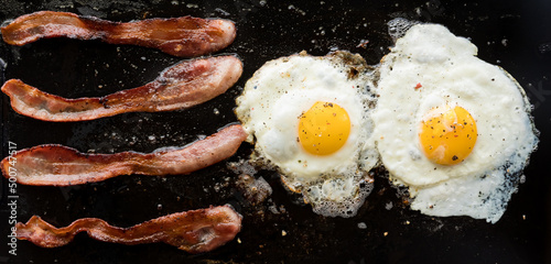 Narrow view of bacon and eggs frying on a cast iron griddle pan.