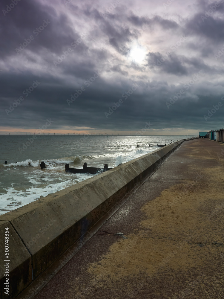 A sweep of sea wall and promenade at Frinton, standing against breaking waves under a stormy sky with a tear admitting sunlight.