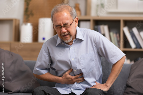 Senior men holding his stomach feeling pain while sitting on the sofa at home. Asian man having stomachache with isolated gastric. Senior suffering from digestion problem or acid reflux. photo